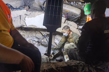 Un soldat portant un uniforme de camouflage, un masque et une lampe frontale parmi les décombres d'une structure effondrée avec d'autres sauveteurs. Il y a une lampe solaire et des bouteilles
