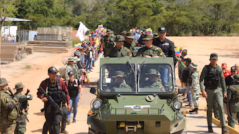 Véhicule militaire blindé vert avec des soldats à l'intérieur et autour, escortés par de l'infanterie armée, parmi une foule avec des drapeaux et à côté de tas de bois