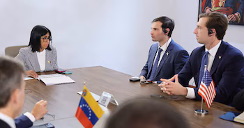Une femme et deux hommes sont assis à une table en bois. Il y a des drapeaux du Venezuela et des États-Unis, des documents et des téléphones