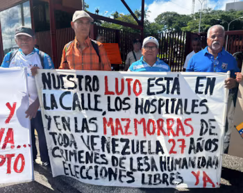 Plusieurs hommes tiennent une banderole blanche avec un texte en noir et rouge qui dit : "NOTRE Deuil EST DANS LA RUE, DANS LES HÔPITAUX ET DANS LES DONJONS DE TOUT LE VENEZUELA 27 ANS DE CRIMES CONTRE L'HUMANITÉ ÉLECTIONS LIBRE MAINTENANT"