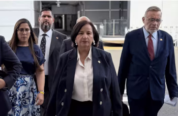 Une femme en costume bleu foncé et chemise blanche au centre, un homme en costume bleu foncé et cravate rouge à droite, et une femme avec des lunettes, une chemise bleue et une jupe imprimée à gauche. Il y a d'autres personnes partiellement visibles. L'arrière-plan est un bâtiment