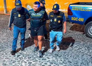 Deux policiers en uniforme et casquettes DEIC escortent un homme en tee-shirt et short. Le groupe marche sur les pavés à côté d’un véhicule de police bleu