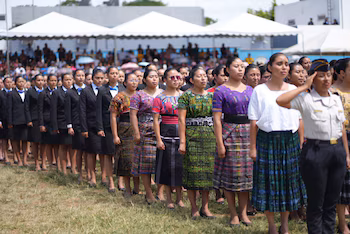 Rangée de femmes dans un champ. Devant, une femme en uniforme salue. Derrière, d’autres portent des costumes noirs avec des nœuds papillon bleus ou des tenues traditionnelles colorées.