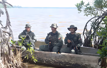 Trois soldats en uniforme, armés de fusils et de chapeaux, voyagent à bord d'un canot le long du rivage d'un plan d'eau recouvert de végétation.