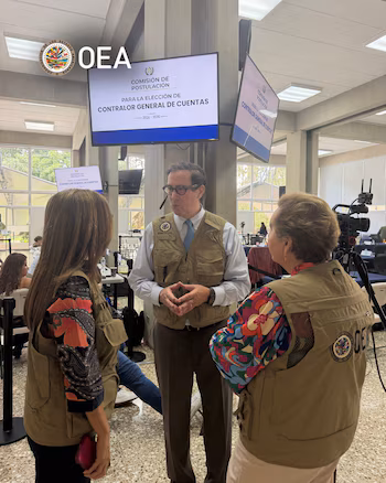Trois personnes portant des gilets avec le logo de l'OEA parlent devant un écran de l'élection du Contrôleur général des comptes