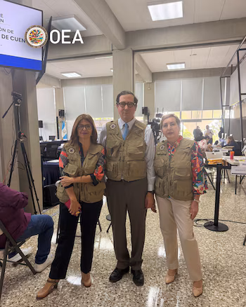 Trois observateurs en gilets beiges posent debout dans une pièce avec des tables, des ordinateurs et le drapeau du Guatemala en arrière-plan