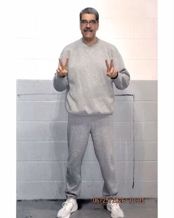 L'homme avec moustache et lunettes porte des vêtements de sport gris et des baskets blanches tout en faisant un geste de victoire avec les deux mains