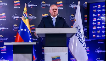 Homme en costume-cravate sur le podium avec le drapeau du Venezuela à sa gauche et le drapeau du ministère de la Justice et de la Paix à sa droite, devant un fond bleu
