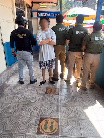 Homme au visage pixelisé, officier du SGIC d'INTERPOL en gilet noir, trois officiers de l'INM en uniforme vert olive. Panneau « MIGRATION ». Sol carrelé, repères de distance