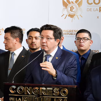 Homme en costume bleu foncé, lunettes, parlant dans un microphone depuis un podium. Derrière, d'autres hommes en costume. Fond blanc avec bouclier et texte "Congrès de la République"