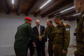 Un homme en costume noir et cravate orange salue un homme en uniforme vert avec un béret rouge, tandis que des soldats en uniforme avec un drapeau israélien le regardent