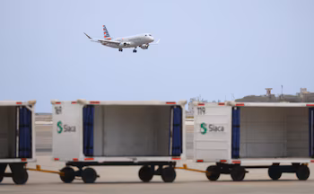 Un avion American Eagle blanc avec une queue rayée rouge, blanche et bleue descend dans un ciel clair, au-dessus de wagons cargo Siaca flous