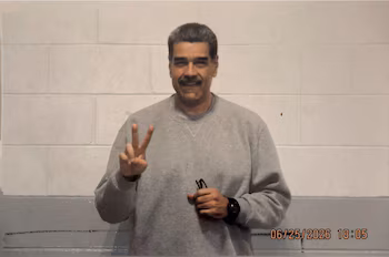 Nicolás Maduro porte un sweat-shirt gris, fait le signe de la victoire avec sa main et tient des lunettes devant un mur de blocs