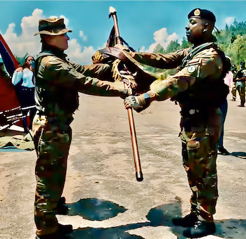 Deux soldats en uniforme de camouflage tiennent un mât en bois avec un drapeau plié dans un espace extérieur