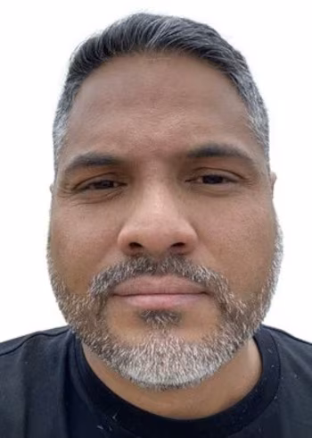 Portrait de face d'un homme aux cheveux gris courts, à la barbe courte et au t-shirt noir sur fond blanc