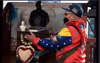 Un homme portant une casquette et une veste colorée parle devant un buste sombre, une composition florale en forme de cœur et une pancarte