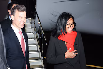 Une femme portant une écharpe rouge, des lunettes et un manteau noir marche à côté d'un homme en costume devant les escaliers d'un avion