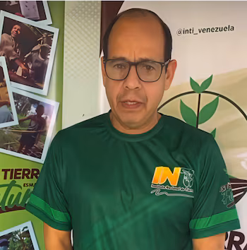 Homme avec des lunettes et un t-shirt vert devant un panneau décoré de photographies et d'un compte de réseau social