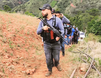 Un homme avec une casquette, un gilet tactique et une arme d'épaule dirige un groupe marchant le long d'un chemin de terre sur une colline