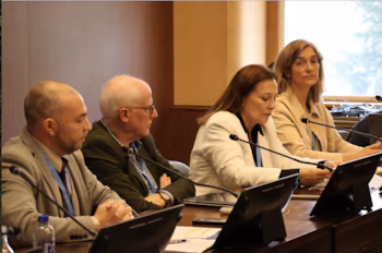 Quatre personnes assises à une table avec des microphones, des moniteurs, des ordinateurs portables et des documents dans une salle de conférence