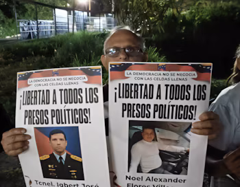 Un homme avec des lunettes tient deux pancartes avec des portraits et le texte Liberté pour tous les prisonniers politiques à côté du drapeau du Venezuela