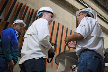 Trois hommes portant des casques de sécurité blancs à l’intérieur d’une installation industrielle. Deux parlent de profil. Un mur avec des rayures colorées verticales