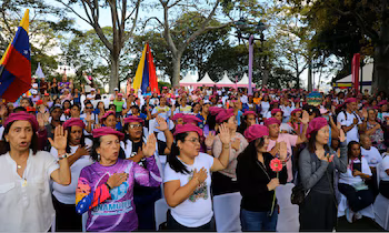 Plusieurs femmes portant des bérets roses lèvent une main ou la posent sur leur poitrine lors d'un événement en plein air avec des drapeaux vénézuéliens en arrière-plan