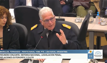 Un homme en uniforme et lunettes parle dans un microphone dans une salle de réunion avec un socle d'information sur l'écran