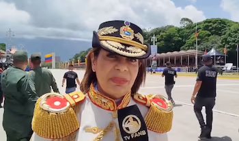 Gros plan d'une militaire en uniforme et casquette avec bouclier, devant un microphone de la FANB TV sur une esplanade