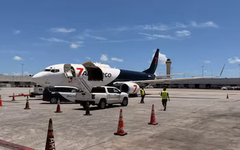 Avion cargo blanc et bleu avec portes ouvertes à l’aéroport. Un camion, une camionnette, des cônes et deux ouvriers sur la piste. Tour de contrôle visible