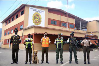 Six personnes en uniforme avec des masques et un chien posent devant un bâtiment avec l'enseigne Commandement national anti-drogue