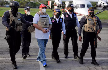 Un homme menotté marche le long d’une piste d’atterrissage entouré d’officiers en uniforme portant des gilets tactiques devant un petit avion
