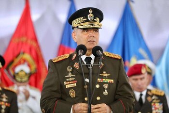 Vladimir Padrino López porte un uniforme militaire avec une casquette et des insignes et parle dans deux microphones avec des drapeaux derrière
