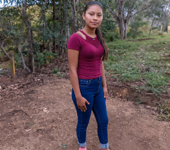 Une jeune femme debout sur un chemin de terre, portant un t-shirt magenta et un jean bleu, avec des arbres en arrière-plan